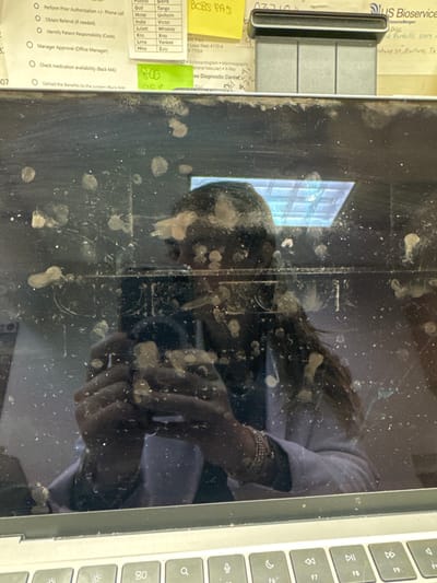 The image displays a person's reflection in a dirty laptop screen, overlaid with visible fingerprints and smudges. The reflection captures the person wearing a white shirt and bracelets, operating the laptop. Adjacent to the screen, a portion of the keyboard is visible. Paperwork with a checklist, possibly related to patient care, and a label with "US Bioservice" are placed beside the laptop. The paperwork includes sections for prior authorization, referrals, patient responsibility, and medication availability. There is a handwritten note on a yellow sticky note.