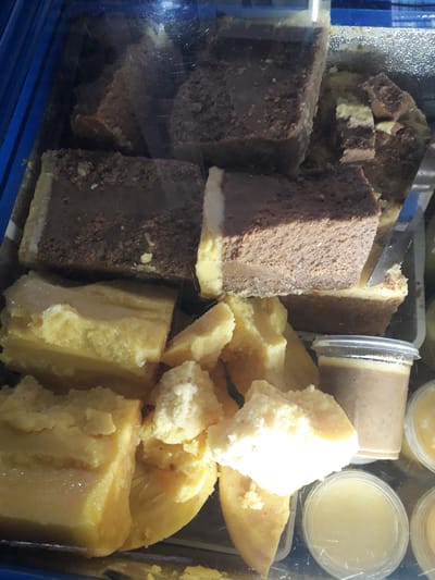This media file captures a close-up view of various sweet food items, likely desserts, displayed within what appears to be a refrigerated case. The scene is brightly lit, revealing the textures and colors of the different treats.

On the left side of the frame, several rectangular blocks of a dark brown, chocolate-colored dessert are prominent. These items exhibit a layered structure, featuring a rich, slightly textured upper layer that contrasts with a smoother, lighter-colored base. Some pieces are whole blocks, while others appear to be cut or broken, suggesting they are ready for serving or have already been partially sold. A metal serving utensil, possibly a knife or spatula, is visible among these chocolate items, reinforcing the idea of a serving station.

To the right, a collection of yellow-colored food items is arranged. These pieces vary in form; some are solid, irregularly shaped chunks or slices with a glossy finish, while others are softer, more crumbly bits of a paler yellow or off-white hue. These could be fruit-based desserts, a type of pudding, or a dense cheese.

In the upper right portion of the image, several small, transparent plastic cups are visible. Each cup contains a smooth, pale yellow, semi-liquid or custard-like substance, individually portioned and sealed, possibly for takeaway.

The overall impression is that of a dessert display in a local market, bakery, or shop in Ghafurov, Tajikistan. There are no people visible, and no discernible text appears on any of the items or packaging. The specific time of day or weather conditions cannot be determined from the image.