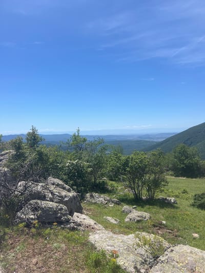 A landscape photograph shows a mountain scene at Marino Pole, Bulgaria, with a clear blue sky. The viewpoint is from a rocky, grassy area, possibly a mountain slope. Vegetation includes trees and grass. In the distance, the landscape extends into a hazy valley with outlines of distant mountains.