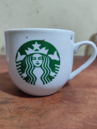 A close-up shot shows a white ceramic mug with the Starbucks logo printed in green on its side. The logo features a siren with flowing hair and a crown. The mug is sitting on a wooden surface, possibly a table, with visible grain and scratches. In the background, a beige-colored wall is out of focus. There are a few small, dark specks on the mug, possibly dirt or coffee residue. The lighting appears to be natural, suggesting it might be daytime. The overall scene is simple and focused on the mug, implying it's a still life composition. No specific activity is depicted, and the setting offers minimal contextual clues beyond the mug and wooden surface. No visible text other than the Starbucks logo. The location context of Lancokpanteara, Indonesia is not visually evident in the image.