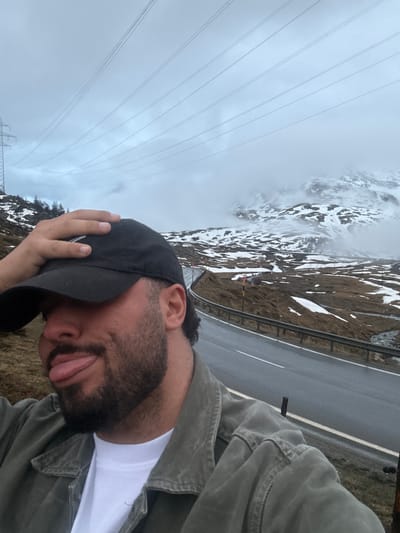 A man takes a selfie outdoors in a mountainous, overcast landscape, likely near Pontresina, Switzerland. The image is taken from a slightly low angle, showing the man in the foreground and a mountain road and cloudy peaks in the background.
The man, positioned on the right side of the frame, is wearing a dark (possibly olive green) jacket over a white t-shirt and a black baseball cap. He has dark hair and a full beard. His head is tilted downwards, and he has a playful expression with his tongue slightly sticking out. His right hand is resting on the brim of his cap.
Behind him, a paved road with a white dashed line runs along the middle distance, bordered by a metal guardrail. The terrain beyond the road is rugged and mountainous, with visible patches of snow covering brown earth and sparse vegetation, suggesting a cold or wintry environment. The distant mountains are partially obscured by thick clouds or mist. Overhead, several parallel power lines stretch across the grey, overcast sky. A power line structure is faintly visible in the lower left background. The overall weather appears cloudy and muted, without direct sunlight. There is no visible text in the image.