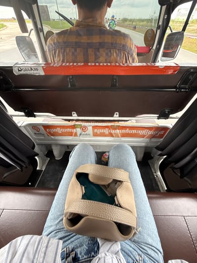 The image depicts the interior of a vehicle, possibly a tuk-tuk, on a street in Krong Ta Khmau, Cambodia. The perspective is from a passenger seated in the rear, looking forward. A person wearing a plaid shirt is driving the vehicle. In the foreground, the legs of another passenger are visible, clad in blue jeans, with a light brown handbag resting on their lap. The vehicle's interior features orange accents with logos and text in Khmer, identifying "Pass App Cambodia Taxi Booking." Outside the vehicle, the environment suggests an urban or suburban setting with roads, some greenery, and a cloudy sky. The scene appears to be captured during daytime.