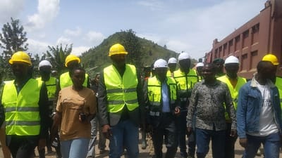 A group of 13 people, primarily men, are walking along a dirt road. Most of them are wearing yellow safety vests and hard hats, some are wearing white hard hats. The majority of the men are African, and one woman is present. The group is walking toward a large, red, articulated dump truck, with its bed raised to reveal its red, dirt-filled contents. There is a lush, green, forested hillside in the background. Banana trees line the dirt road, and the sky is a bright blue with white clouds. The scene appears to be a construction site in Sake, Democratic Republic of Congo, with coordinates (latitude: -1.57, longitude: 29.045).