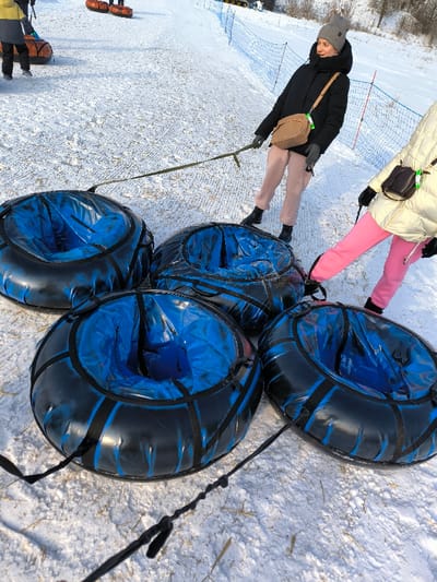 The scene depicts a snow-covered slope at a recreational facility in Prybuzke, Ukraine, under clear daylight conditions. Four black and blue snow tubing inner tubes are arranged in the foreground on the snow. Each tube has a visible blue interior and black exterior with black webbing and handles. Two female individuals are present, standing near the tubes. One woman, positioned centrally, wears a gray knitted beanie, a black puffer jacket, pink pants, black boots, and gray gloves; she has a tan cross-body bag and holds a long green strap connected to the lead tube. The second person, partially visible on the right, wears a light-colored puffer jacket, pink pants, and black boots; they have a dark cross-body bag and their right foot is near one of the tubes, with a black strap extending from it. Both individuals appear to be preparing or pulling the connected snow tubes uphill. In the background, a blue safety net runs along the right side of the slope, and other individuals on orange snow tubes are visible further up the incline.
