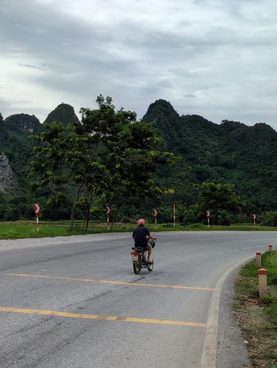 A single person, wearing a dark shirt and shorts, is riding a moped down a paved road. The rider has a pink helmet on and is facing away from the camera. The road has yellow lines painted on it, indicating a crosswalk or junction. In the background, there are lush green mountains with a cloudy sky. There are also trees and roadside markers visible. The overall scene appears to be in a rural or semi-rural area. The location is Tân Thanh Commune, Vietnam.