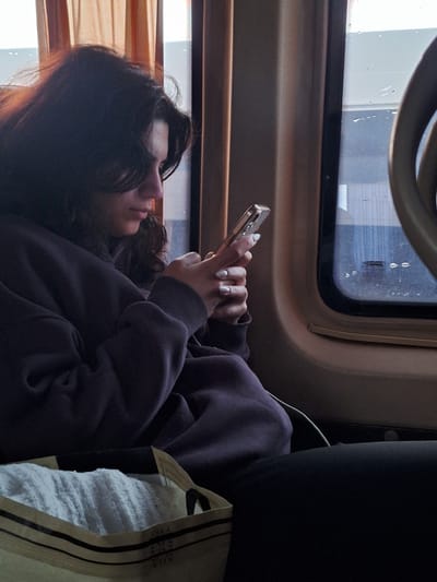A young woman is seated on a train, looking at her phone. She has long, dark curly hair and is wearing a dark purple sweatshirt. Her hands are holding a gold-colored smartphone. To her right is a white bag with dark stripes. Behind her is a black headrest. The train is traveling past windows with a view of buildings. The image provides no information about the city of Anipemza, Armenia.
