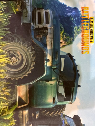 The image is a close-up, vertically oriented photograph of a portion of a printed surface, likely a mousepad or fabric. The central subject is the front quarter of a teal-colored truck, reminiscent of a military vehicle or heavy-duty off-road vehicle. Its large, lugged tire is prominently featured on the left side of the frame, with detailed tread patterns visible. The truck's body is painted a metallic teal, with a silver-colored hood and front grille assembly.
To the right of the truck, vertical text in bold, yellow and orange lettering reads "BATTLEGROUNDS" and below that "PLAYERUNKNOWN'S." This text is aligned with the side of the truck and is part of the printed design.
The background of the printed image depicts a lush, green, mountainous landscape with trees and foliage under a cloudy sky. The foreground, surrounding the truck, features out-of-focus green and yellow foliage, suggesting a natural, outdoor setting.
The photograph itself is taken indoors, with the printed surface laid flat. The lighting is natural, casting soft shadows. There are no people present in the image, and no actions or interactions are depicted. The overall scene is a still, printed graphic. The image does not contain any information that would allow for its location in Melitopol, Ukraine, or any other specific city.