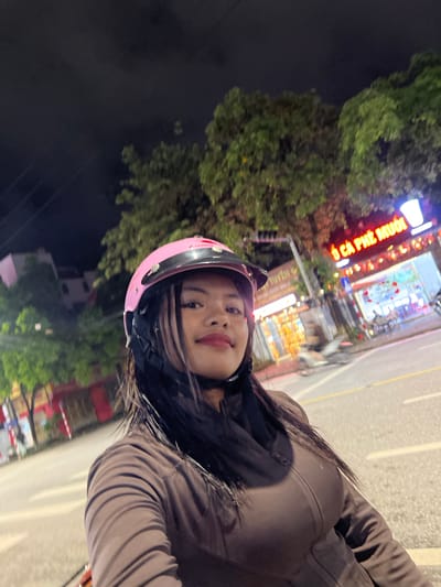 A young woman wearing a pink helmet and a brown jacket is taking a selfie at night in Minh Xuân Ward, Vietnam. The background shows a street with trees, buildings, and neon signs, indicating an urban setting. One sign reads "CÀ PHÊ MUỐI" in large, illuminated letters, suggesting a coffee shop. The road appears wet, possibly from rain. The overall scene depicts a casual moment captured during nighttime in a Vietnamese city.