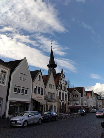 The image depicts a daytime street scene in Burgsteinfurt, Germany. A long row of multi-story buildings with varied facades lines a cobblestone street. Dominating the upper right-center is a historic building featuring a tall, dark, pointed spire with a weathervane and a visible clock face. Adjacent to it on the left is a prominent white building with ornate arched windows, decorative elements, and a red-tiled gabled roof. Other buildings in the row have gabled roofs and varying exterior colors, from light grey to white and beige.
On the left side of the street, a silver Hyundai i20 hatchback (German license plate ST H 6064) is parked on the cobblestones in front of a building housing "Bäckerei Drobisch Konditorei" and "Eiscafé Di Marco." Another dark sedan is parked directly behind the Hyundai. Further down the street on the right, several other vehicles are present, including a dark sedan and a silver sedan (German license plate MS TS 703), as well as a white van.
In the mid-ground, numerous empty outdoor tables and chairs are arranged on the cobblestones in front of the businesses, some with large, closed umbrellas. No individuals are clearly visible. The sky above is blue with scattered white stratocumulus clouds. The lighting indicates daylight conditions.