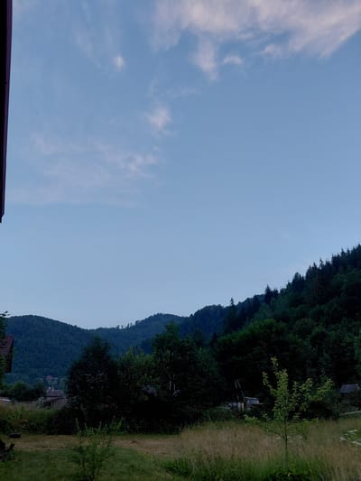 The image depicts a rural landscape with rolling hills covered in dense green forests under a pale blue sky with wispy clouds. In the foreground, a grassy area is visible, with some scattered vegetation. To the left, part of a building with a dark roof is visible. Further back, a small cluster of houses and structures can be seen nestled amongst the trees on the slopes of the hills. The overall impression is of a tranquil, natural setting, likely in a village or countryside location. Given the prompt mentioning Chernyk, Ukraine, this scene could be representative of the rural outskirts of such a location, characterized by its mountainous terrain and prevalent forestry. There are no people or animals visible in the image, and no discernable actions or interactions are taking place.
