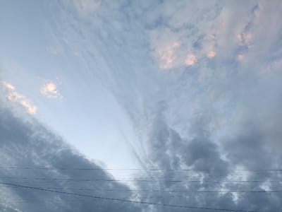 The image captures a wide-angle view of the sky above Kyzyl-Suu, Kyrgyzstan. The sky is a blend of soft blues and grays, with scattered clouds. Some of the clouds exhibit a subtle pinkish hue, suggesting the influence of dawn or dusk light. Multiple thin, dark wires stretch horizontally across the lower portion of the frame, indicating the presence of overhead power lines. These wires are distinct against the lighter sky and darker clouds, adding a man-made element to the natural scene. The overall impression is one of a serene, open sky with the subtle interruption of utility infrastructure.