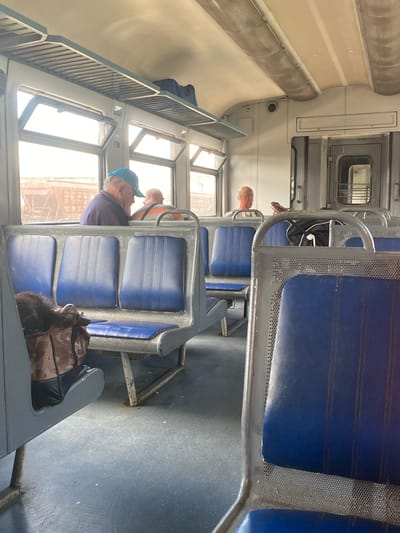 An interior view of a train carriage shows rows of blue-padded seats with metal frames. In the background, three men are visible, two seated and one standing, looking out the window. The man in the foreground, wearing a blue cap, is facing away from the camera. The train carriage is clean and well-lit, with luggage racks above the seats. The overall impression is one of a typical public transport setting. The photo was taken in Romodan, Ukraine.