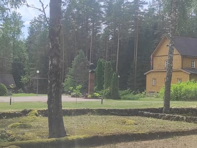 In the city of Litvenski Sel'ski Sovet, Belarus, a scene unfolds with a yellow wooden building to the right, featuring a dark roof and several windows. In front of the building, a tall, dark tree stands prominently. A stone monument, possibly a bust on a pedestal, is visible in the middle ground, flanked by several evergreen trees. The ground is covered in grass, with some areas of mossy stones in the foreground, suggesting a park or memorial setting. A paved path runs through the grassy area. The background is dominated by a dense forest of pine trees.