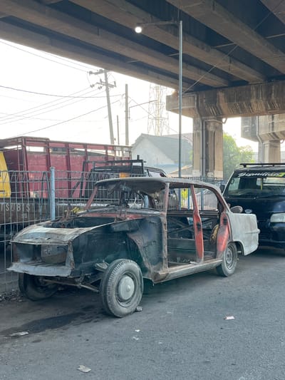 A heavily damaged and stripped-down car body sits on a paved road under an elevated concrete structure. The car, which appears to be a sedan with its doors open and most of its interior removed, is rusted and appears to be in a state of disrepair. A black vehicle with the word "Bismillah" on its windshield is parked to the right of the damaged car. In the background, a red truck trailer with a metal fence in front of it is visible, along with utility poles and buildings. The scene is likely located in Tenggulunan, Indonesia, as indicated by the presence of local infrastructure and vehicles. The lighting suggests it is daytime, with the sky overcast.