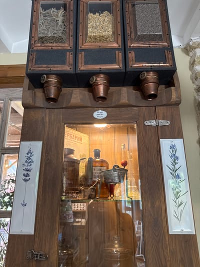 In Semogorskiy, Russia, a vintage wooden apothecary cabinet stands against a stone wall. The upper section features three glass-fronted compartments filled with dried herbs and seeds, each with a copper dispensing mechanism below. The main body of the cabinet is a wooden case with a glass door, revealing shelves stocked with antique medicinal bottles, a copper jug, a tin labeled "MOULINETTE," and other assorted glass containers. Two decorative ceramic tiles, one on each side of the cabinet door, depict sprigs of lavender. The lighting suggests an interior setting, possibly a shop or a historical display.