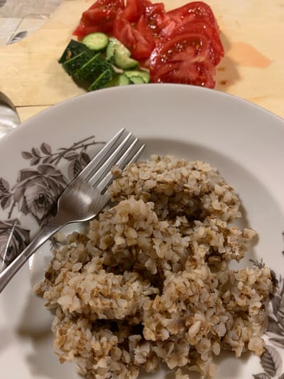 The image presents an overhead, close-up view of a prepared meal set on a light-colored wooden surface, possibly a cutting board or table. The central subject is a round, white plate containing a generous portion of cooked buckwheat. A silver, four-pronged fork rests across the plate, with its handle extending towards the bottom right of the frame. The plate is adorned with a detailed grey and black floral pattern, depicting roses and leaves, visible around the edges and beneath the fork.

To the left of the plate, on the wooden surface, is a fresh side salad composed of sliced vegetables. There are vibrant red tomato slices, appearing juicy, and green cucumber pieces, some cut into rounds and others into short, rectangular segments. A small amount of reddish liquid, likely tomato juice, is visible on the wooden surface beneath the vegetables.

The scene depicts a simple, home-style meal, ready to be eaten. The lighting is soft and even, suggesting an indoor setting. No people or visible text are present in the image. The food items, particularly the buckwheat, align with culinary traditions found in the given location context of Kasimovo-1, Russia.