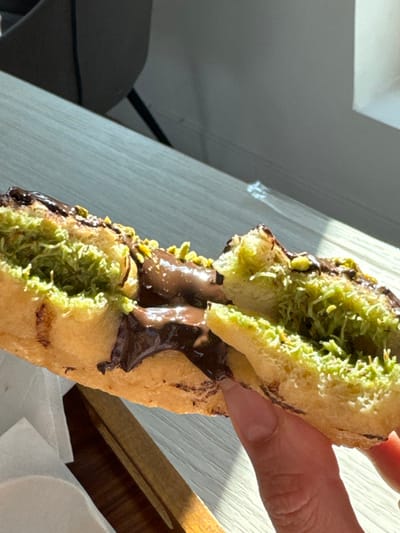 A close-up image depicts a pastry, possibly a cronut or similar fried dough, held by a hand. The pastry is split lengthwise, revealing a rich, dark chocolate filling that is thick and slightly molten, oozing from the center. The exposed inner surfaces of the pastry are coated with a vibrant, shredded green substance, possibly coconut or pistachio, with a slightly fibrous texture. The exterior of the pastry is a golden brown, with dark chocolate drizzle and scattered small, light-green chopped nuts (likely pistachios) visible on its top surface.
A light-skinned hand, with neatly manicured nails, holds the pastry from the underside, near the split. In the foreground to the left, a portion of a white napkin is visible. The pastry rests partially over a light-colored wooden surface, possibly a table or tray. Strong, natural sunlight illuminates the scene from the right, creating distinct shadows on the wooden surface and highlighting textures. The background is out of focus, showing blurred shapes consistent with an indoor setting, including a dark, curved object that could be part of a chair and a light-colored wall. The scene is located in San Phra Net, Thailand.