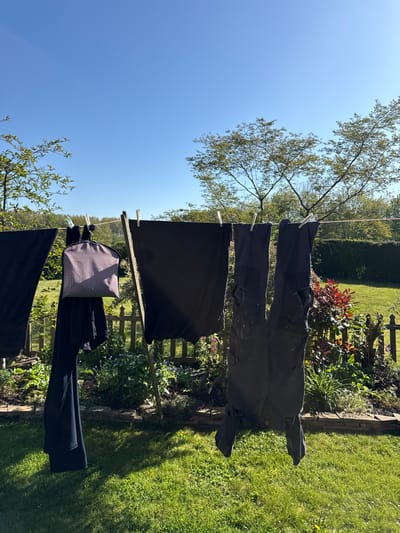 The image displays a bright, sunlit outdoor scene of laundry drying on a clothesline in a garden, located in Hitchin, United Kingdom. The perspective is slightly upward, with the clothesline stretched across the midground, holding several dark-colored garments.

Hanging on the line are multiple items, secured by white clothespins. These include at least two pairs of dark trousers or work pants, some showing faint lighter marks or wear. A large piece of dark fabric, possibly a sheet or duvet cover, is draped over the line. Further along, a distinct black and white houndstooth patterned item, appearing to be a cushion cover or small bag, is visible next to another dark garment.

Above the clothesline, a wide expanse of clear, vibrant blue sky fills the upper part of the frame, with green leafy tree branches extending into the upper left and right corners. Below the clothesline, a well-tended green lawn features prominent, elongated shadows, indicative of strong sunlight. Along the right side of the frame, a garden bed thrives with various plants and foliage, including some with reddish hues, bordered by a rustic wooden fence or trellis. Dense green hedges are visible further in the background, adding to the garden setting.

The scene captures a peaceful, domestic activity, with clothes being air-dried on a clear, sunny day, likely in the morning or afternoon given the distinct shadows. No people or visible text are present in the image.