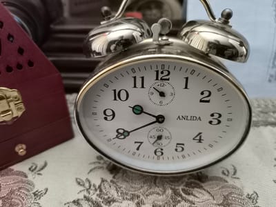 The image is a close-up, eye-level shot of a vintage-style, silver-colored analog alarm clock. The clock face is white with black Arabic numerals marking the hours. A smaller dial within the main face indicates seconds. The hour hand points to just after 9, and the minute hand points to approximately 47. The clock has two bells on top, characteristic of an alarm clock. To the left of the clock is a portion of a dark red, ornate wooden box with gold-colored hardware, possibly a jewelry box or keepsake box. The objects are placed on a white tablecloth with an intricate, grey floral pattern. The background is blurred, suggesting a shallow depth of field, and appears to be composed of various indistinct objects and textures. The scene does not provide any information about the city of Dolakovo, Russia, or any individuals.