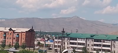 The image shows a panoramic view of a city with a mountain range in the background. In the foreground, there are several residential buildings, including a red brick building on the left and a multi-story building with a green roof on the right. There are also utility poles and some trees visible. In the mid-ground, there are smaller buildings and a body of water. The mountain range in the background is barren and appears to be covered in snow in some areas. There are some clouds in the sky.

The image is likely taken in the city of Kyzyl, Russia, given the presence of typical Soviet-era apartment buildings and the surrounding landscape, which is characteristic of the region. The specific location within Kyzyl is not identifiable from the image alone, but it appears to be a residential area on the outskirts of the city, with views of the mountains.