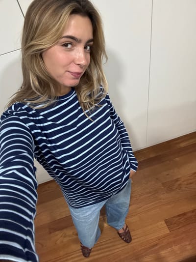 A nearly full-length, high-angle selfie photograph captures a female subject standing indoors, in a location identified as Cidade da Maia, Portugal. The woman has long, wavy blonde hair, parted on the side, and is looking directly at the camera with a subtle smile. She is dressed in a long-sleeved navy blue shirt with light blue horizontal stripes, light-wash denim jeans, and dark brown woven closed-toe flat shoes. Her left arm, extended, holds the camera, and her right hand is casually placed in her right jeans pocket. The immediate background features a plain, light beige wall with visible vertical seams, suggesting panels or cabinetry. The floor is made of polished light brown wooden planks.