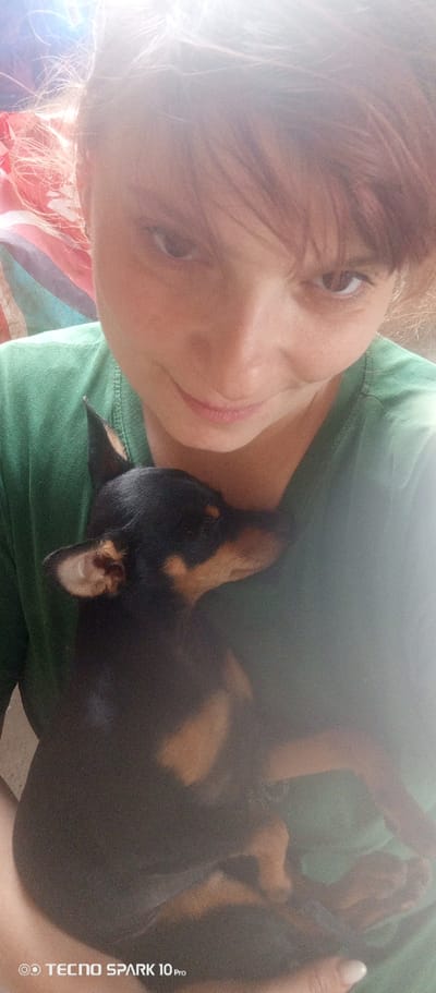 A woman is holding a small black and tan dog in her arms. She is wearing a green t-shirt. The dog is positioned against her chest. The picture is taken in an indoor setting. The bottom left corner has the text "TECNO SPARK 10 Pro". The location is Bereg Kubani, Russia.