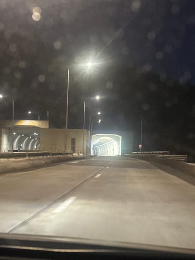 The image shows a tunnel entrance illuminated at night. Streetlights line the roadway leading to the tunnel. A large, concrete wall runs along the right side, with a vertical vent system visible at the top. The tunnel's interior is lit, revealing a curved, arched structure. A red light hangs from a pole above the tunnel entrance. Raindrops are visible on the window, blurring the left side of the image.