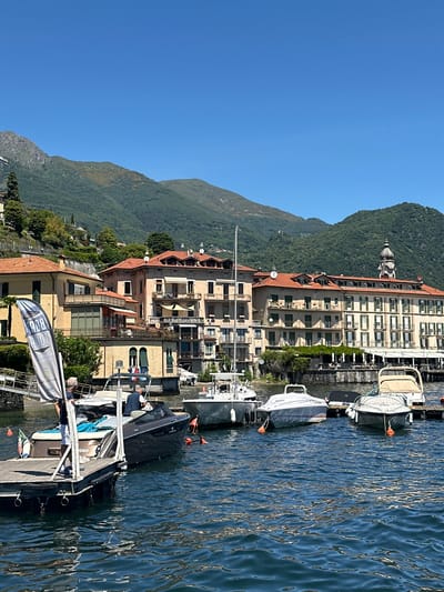 The image depicts a sunny day in Loveno, Italy, with a view of boats docked on a clear blue lake. In the foreground, several motorboats are moored to a wooden dock. One person stands on the dock next to a dark-colored boat, holding a flag. In the background, a row of buildings with terracotta roofs lines the shore, and lush green mountains rise behind them. The architecture suggests a European lakeside town. The water is calm, reflecting the clear sky and the surrounding scenery.