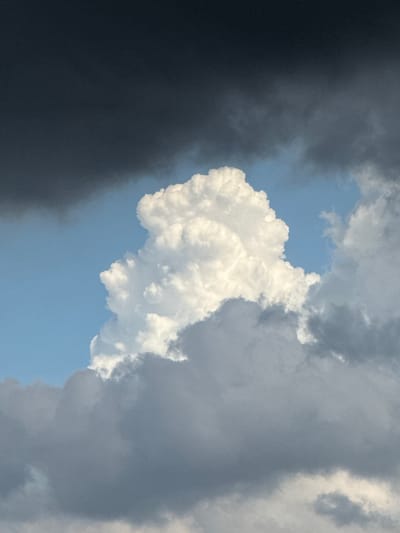 The image depicts a sky with dramatic cloud formations, located in Đa Lộc, Vietnam. The sky is a mix of deep charcoal gray clouds in the upper left and lighter, turbulent gray clouds lower down and to the right. A prominent, brightly lit cumulus cloud, appearing white and puffy, rises centrally against a bright blue sky, suggesting a break in the stormy weather. The contrast between the dark and light clouds creates a dynamic and somewhat ominous atmosphere. There are no people or man-made objects visible in the frame, focusing solely on the natural phenomenon of the clouds. The scene is entirely environmental.