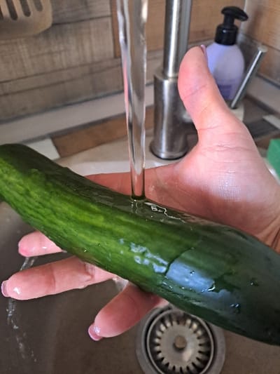 A kitchen sink area, located in the city of Yegoryevsk, Russia, is depicted. A human left hand, with light pink polished fingernails, holds a dark green, ribbed cucumber horizontally. A continuous stream of clear water flows downwards from a chrome or stainless steel kitchen faucet, impacting the top surface of the cucumber. The cucumber and the visible portions of the hand are wet. Below the hand, a metallic sink basin with a circular drain stopper assembly is partially visible. The background features a tiled backsplash with rectangular tiles exhibiting beige and brown tones. To the right of the faucet, a purple bottle with a black pump dispenser, likely liquid soap, is present, alongside a partial view of a green rectangular sponge.