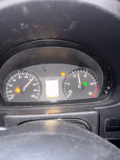 The image shows a close-up view of a car's dashboard, specifically the instrument cluster. The speedometer on the left indicates a speed of approximately 70 km/h. To the right of the speedometer is a tachometer, showing engine RPMs multiplied by 100, with the needle pointing slightly above 10 (indicating around 1000 RPM). A digital display between the two gauges shows a fuel level that appears to be nearly full. There are two warning lights illuminated: an orange engine light on the speedometer and a green light on the tachometer. The dashboard is dusty. The image is taken inside a vehicle, and there is no indication of the city of Ornaisons, France, or any external environmental context.