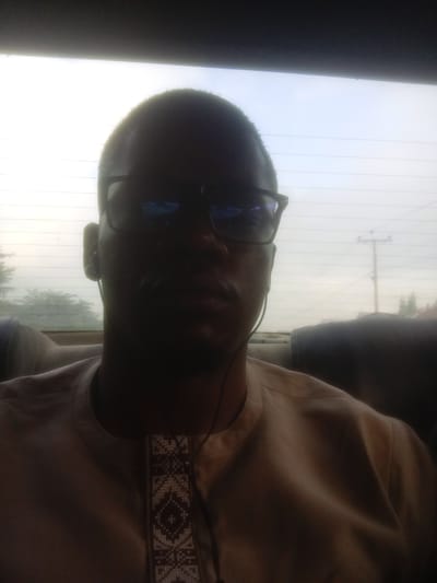 A man wearing glasses and a beige shirt with a patterned neckline is pictured from the chest up. He is sitting in the back seat of a car, with the sky visible through the rear window. The lighting suggests it is daytime, possibly overcast. There is a power pole and trees visible in the background outside the car. The image is slightly blurry. The location is identified as Sudbury, United States of America.