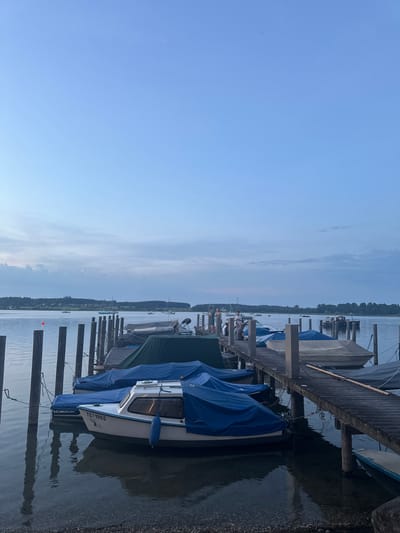 The image depicts a dusk scene at a marina on the Chiemsee lake in Rimsting, Germany. Wooden piers extend into the calm water, where several boats are moored. Many of the boats are covered with blue tarpaulins, providing protection from the elements. One white boat in the foreground has the registration "TS-1965" visible. On one of the piers, a small group of people are standing and sitting, appearing to be socializing. The sky is a pale blue with scattered clouds, transitioning from daylight to evening. The shoreline in the background is lined with trees and hints of houses, suggesting a lakeside town or village. The water reflects the sky and the piers, creating a serene atmosphere.
