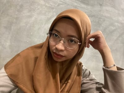 A young woman wearing a brown hijab and silver-rimmed glasses is pictured in a selfie taken in Sekar Mawar, Indonesia. She is looking to her left, with her right hand resting on her head. She is wearing a light-colored, possibly corduroy, collared shirt. The background is a textured, gray wall.