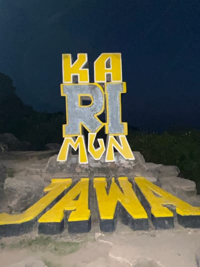 The image displays a large, prominent sign with the words "KARIMUN JAWA" in bold, three-dimensional lettering. The letters are primarily yellow with white and gray accents, and they are mounted on a rough, stone-like base. The sign appears to be a landmark or welcome sign for the location of Karimunjawa, an archipelago in Indonesia known for its marine national park. The background is dark, suggesting it is nighttime or dusk, with the sky a deep blue-gray. There are subtle indications of foliage and rocky terrain in the background, consistent with a natural or semi-natural setting. No people are visible in the image.