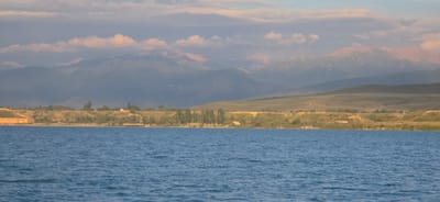 This image depicts a serene lake with a mountainous backdrop. In the foreground, the deep blue water of Issyk-Kul Lake is visible, with gentle ripples disturbing its surface. The middle ground features a shoreline lined with trees and vegetation, interspersed with some structures. On the left side of the shoreline, a long, low building with a light-colored facade is prominent. Further along the shore, a wooden pier extends into the water. Beyond the shoreline, rolling hills lead up to the imposing mountain range that dominates the background. The mountains are layered in shades of blue and grey, with some peaks catching the soft light of what appears to be late afternoon or early morning, casting pink hues on some of the higher elevations. The sky above is a soft pastel, with scattered clouds. The scene is tranquil and appears to be a natural landscape with some signs of human presence near the lake. This location is within or near the city of Pristan'-Przheval'sk, Kyrgyzstan, on the eastern shore of Issyk-Kul Lake.