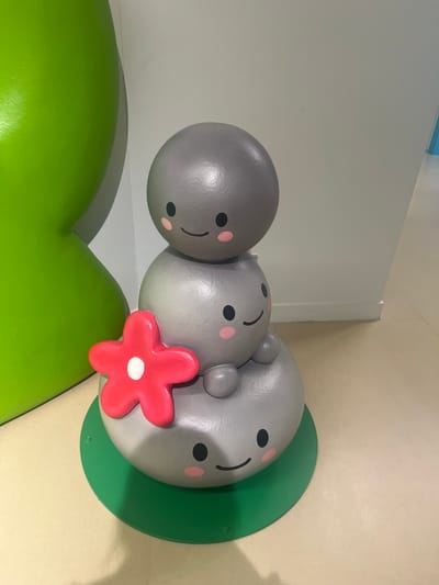 A whimsical sculpture of three stacked gray rocks, each with simple cartoon faces, stands on a green circular base. The top rock has eyes and a smile. The middle rock has eyes and rosy cheeks. The bottom rock has eyes, rosy cheeks, and a gentle smile. A bright red flower with a white center is attached to the side of the middle rock. To the left of the sculpture is a large, bright green, amorphous shape. The background consists of a light-colored wall and floor. This sculpture is located at the entrance of the Dinosaur Museum in Hwaseong-si, South Korea, which is part of the Hwaseong Dinosaur University Town.