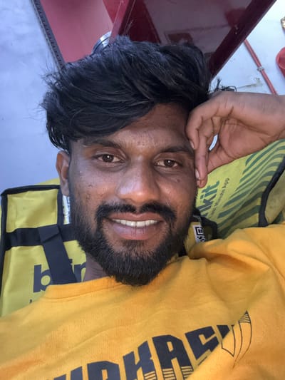 The scene is set in Palghar, India, featuring a dark-skinned adult male positioned in a close-up, chest-up view. He has a dark, full beard, a mustache, and dark, medium-length hair styled to fall across his forehead. His eyes are dark, and he exhibits a broad smile, revealing his upper teeth. His left hand is raised, with fingers resting against his temple area, partially covering the upper right side of his face. He is wearing a plain yellow long-sleeved crew-neck shirt; a dark, partial graphic resembling "ARP" and an outline of pencils is visible on the lower front of the shirt. Behind his left shoulder, a yellow delivery bag with black straps is visible, displaying a partial black "b" logo. Behind his right shoulder, a green and yellow striped object, consistent with a uniform or delivery bag, is visible, bearing white text that reads "KIT" and "Last Minute App". The background includes a light blue-grey surface on the upper left, a dark red/maroon structural element above his head, and segments of a white wall or surface with a thin red cord running diagonally on the upper right. A metallic railing or serrated edge is discernible in the upper left background.
