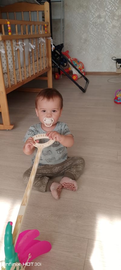 An indoor scene shows a baby sitting on a light-colored wood-effect floor. The baby, with dark hair and brown eyes, is wearing a light blue t-shirt with a print of vehicles and patterned olive green pants, as well as pink socks with orange grips. A white pacifier is in the baby's mouth, and the baby is holding a toy that resembles a rake or a handle with a large attachment. To the left of the baby is a wooden crib with a mattress that has a star pattern. In the background, to the right of the crib, is a part of a playard with red and yellow accents and a black frame, as well as other toys. The wall in the background is covered with patterned wallpaper. There are no visible indicators to place this scene in the city of Kedrovy, Russia.