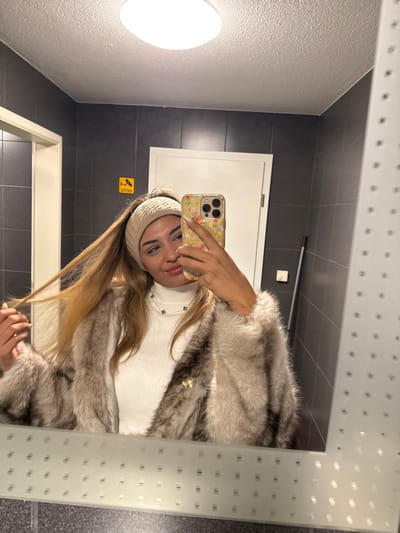 The depicted scene, understood to be located in Bad Vilbel, Germany, shows a female subject captured in a mirror selfie within an indoor environment. The subject, with long, light brown hair, wears a beige knit headband, a white ribbed turtleneck sweater, and a light-colored fur or faux fur coat featuring a small gold-toned emblem on the right lapel area. She also wears a white beaded necklace interspersed with turquoise beads. Her right hand, holding a smartphone with a floral-patterned case, is positioned to take the photograph, partially obscuring the lower right portion of her face. Her left hand is actively manipulating strands of her hair on the left side of her head.
The background, reflected in the mirror, features dark grey tiled walls and a white, textured ceiling with a large, oval-shaped flush-mount light fixture. Two white doors are visible, flanking a yellow square sign with a black pictogram and indistinct letters, affixed to the dark grey wall. A dark-colored handle, possibly from a cleaning implement, leans against the right wall segment, above a white electrical outlet or switch. The mirror's frame exhibits a patterned border of translucent squares along its visible bottom and right edges.