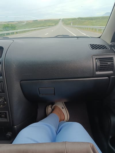 A first-person perspective shot captures the interior of a vehicle as it travels down a highway. The view is dominated by the dashboard, with the windshield showing a blurred landscape of fields and a road stretching into the distance. A crack is visible on the windshield. A person's legs and feet are visible in the lower part of the frame, with light blue pants and white sneakers. The dashboard features an air vent and a glove compartment. The overall impression is one of travel on a clear day in a rural setting, possibly on the outskirts of Utrennyaya Dolina, Russia, given the context.