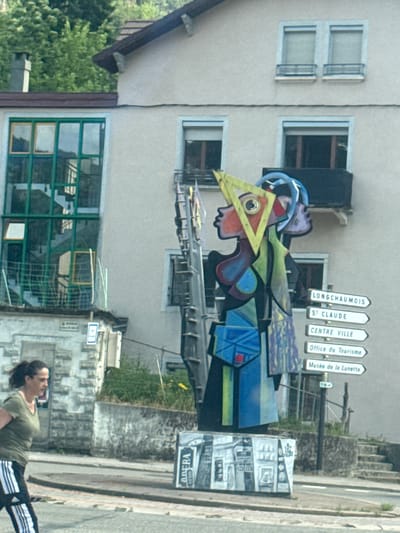 This image captures a street scene in the city of Hauts de Bienne, France, featuring a striking abstract sculpture as the central subject. The sculpture is colorful and geometric, with distinct sections in shades of yellow, red, blue, and green, and appears to be a representation of a stylized human figure. It is mounted on a base that is painted with black and white renderings of buildings.
To the right of the sculpture, a series of white directional signs on black posts point towards various locations: "LONGCHAU MOIS," "ST CLAUDE," "CENTRE VILLE," "Office du Tourisme," and "Musée de la Lunette."
In the background, a building with a pale facade is visible, featuring multiple windows, some with dark frames and others with green frames. A green staircase is visible through one of the large green-framed windows.
On the left side of the image, a woman with dark hair pulled back walks from left to right. She is wearing a green t-shirt and black athletic pants with white stripes. She is in motion, suggesting she is walking or jogging. Adjacent to her is a stone building with a textured facade and a yellow and blue sign.
The overall environment appears to be an urban or semi-urban setting, with a paved road in the foreground and some greenery visible in the background. The sky is not clearly visible, but the lighting suggests it is daytime.