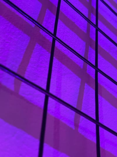 The image displays an abstract, close-up view of a dark, grid-like linear structure, likely composed of metal bars or wires, positioned in the foreground. This structure casts distinct shadows onto a textured background surface, possibly a wall, which is uniformly illuminated by intense, artificial purple light. The shadows, varying in shade from lighter to darker purple, form geometric patterns of alternating diagonal and horizontal stripes on the background. No people are visible. The scene is static, devoid of any discernible action or interaction. The architectural style, use of strong colored lighting, and geometric patterns are consistent with contemporary public or cultural buildings. The location is identified as **Derby Quad**, Derby, United Kingdom.