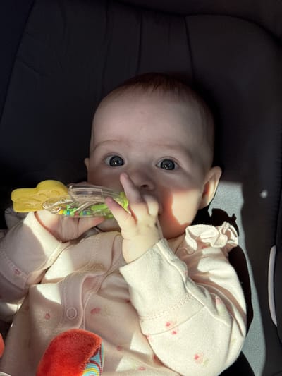 A close-up shot shows a baby girl with blue eyes and fair skin, wearing a light pink pajama set with a floral pattern. The baby is holding a yellow and clear plastic rattle in her mouth and her right hand. A patch of sunlight illuminates her face, creating a shadow on the right side of her head. The background is a dark, textured surface, possibly a car seat or stroller. The image is taken from a high angle, looking down at the baby. The setting appears to be indoors.