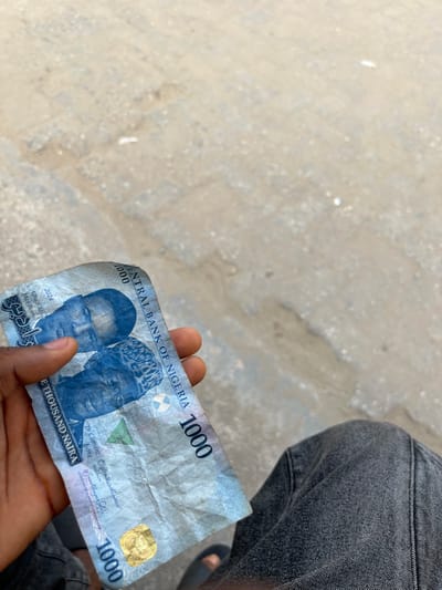 A slightly wrinkled one thousand Naira note from Nigeria is held in someone's hand. The note features portraits of two men, one in a western suit and the other in traditional attire. "Central Bank of Nigeria" is printed above the faces, with the denomination "1000" displayed in multiple locations. The note includes the signatures of Dr. Clement Isong and another individual. The year "2024" and Arabic script are also visible on the bill. The person holding the note wears gray jeans. A gray cement surface is in the background.