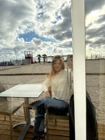 A female subject with shoulder-length blonde hair is depicted in a reflection taking a self-portrait with a smartphone. She is seated at a white rectangular table inside a building with a wooden floor. She wears an off-white V-neck long-sleeve top, distressed blue jeans with knee rips, and dark sneakers. Her left arm rests on the table, and her right hand holds an iPhone with a light-colored, floral-patterned case, from which a black and green braided lanyard hangs. A dark garment is draped over a chair to her right. Visible through the glass window is a sandy beach under a partially overcast sky featuring substantial cloud cover and patches of blue. The beach environment includes a prominent red metal lifeguard tower on the left, multiple wooden-framed beach establishments, two palm trees, and white boundary fencing. The sea is discernible in the far distance on the right, with several indistinct figures present near the waterline. This scene is located in Quartu Sant'Aleni/Quartu Sant'Elena, Italy.