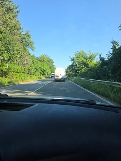 The image is a forward-facing view from inside a vehicle on a two-lane road bordered by lush green trees and vegetation. The sky is clear and blue. In the distance, two white vans are traveling in the same direction. The closest van is a large box van, and behind it is a smaller van. The road has a white line marking the edge and a dashed white line dividing the lanes. The foreground shows the dark dashboard of the vehicle from which the photo was taken. The scene appears to be a rural or suburban setting. The location is identified as Blatino, Bulgaria.
