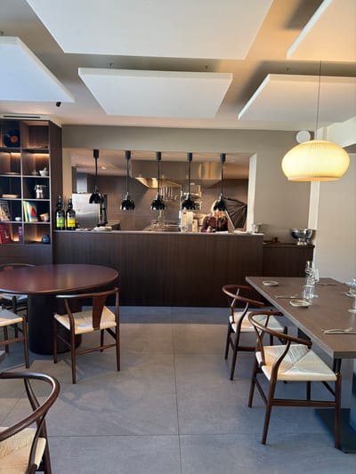 The image depicts the interior of a modern restaurant in Marche-lez-Écaussinnes, Belgium. The well-lit dining area features a large round dark wood table with three dark wood Wishbone chairs, each possessing a light woven seat, positioned on the left. To the right, a rectangular dark wood table is set with multiple place settings, including white plates, glassware, and cutlery, accompanied by additional Wishbone chairs. The floor is composed of large, light gray square tiles.

A dark wood counter with four black pendant lights separates the dining space from an open kitchen visible through a rectangular cutout. Inside the stainless steel kitchen, a single individual with long blonde hair, wearing a dark top, is partially visible, positioned behind the counter and seemingly engaged with an item. The kitchen's rear wall shows various hanging utensils.

A tall dark wood shelving unit on the far left holds diverse objects such as books, a black vinyl record, and ceramic items. The ceiling features several large, rectangular white acoustic panels and recessed lighting, along with a prominent spherical ribbed pendant lamp in the right section of the dining room.