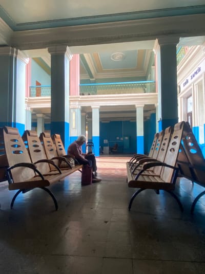 The image shows an elderly person sitting alone on a wooden bench in a waiting area, likely a train station or public building, in Romodan, Ukraine. The person is dressed in a long-sleeved shirt and is looking down, appearing contemplative or tired. Beside them, on the bench, is a patterned bag. On the floor, there is a maroon bag or suitcase. The waiting area features multiple rows of similar wooden benches with unique curved backs and oval cutouts. The architecture is characterized by tall, blue and pink columns, balconies with decorative railings, and a multi-colored ceiling. Natural light streams in from a large opening, casting shadows on the polished floor. The overall atmosphere is one of quiet solitude and waiting.