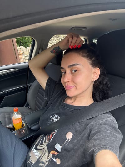 A woman is sitting in the driver's seat of a car. She has dark, curly hair pulled back and is wearing a dark t-shirt with a graphic print. Her right arm is raised, with her hand touching the top of her head, showing red nail polish. She has a tattoo of a butterfly on her left arm. A seatbelt is across her chest. A bottle of orange beverage is in the car's cup holder. The interior of the car appears to be dark-colored. Outside the car window, a stone wall and greenery are visible. The lighting suggests it is daytime. The context of Manavi, Georgia is not discernible from the visual information provided in the image.